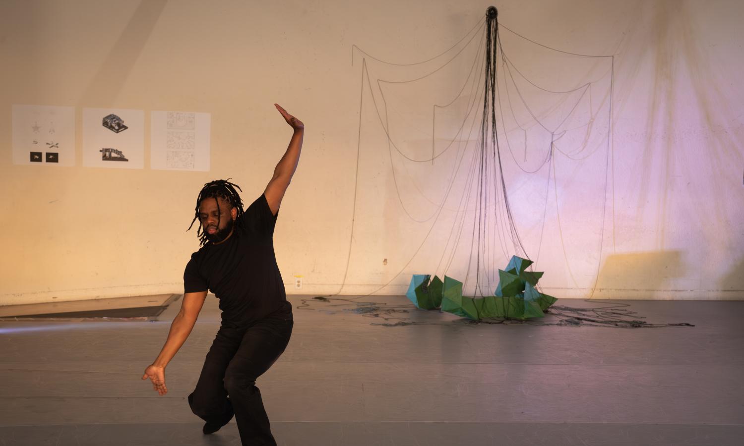 Black non-binary person with locs is wearing black and dancing. Their arms are on an angle and they are in a semi-crouched position.