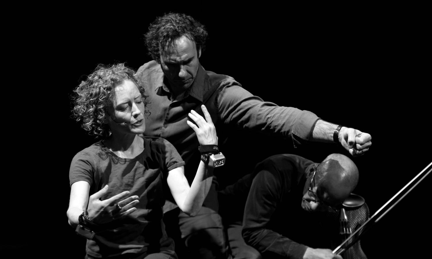 A black and white photograph of three figures against a black background.  Two are gesturing together with their arms.  The third is playing a violin.