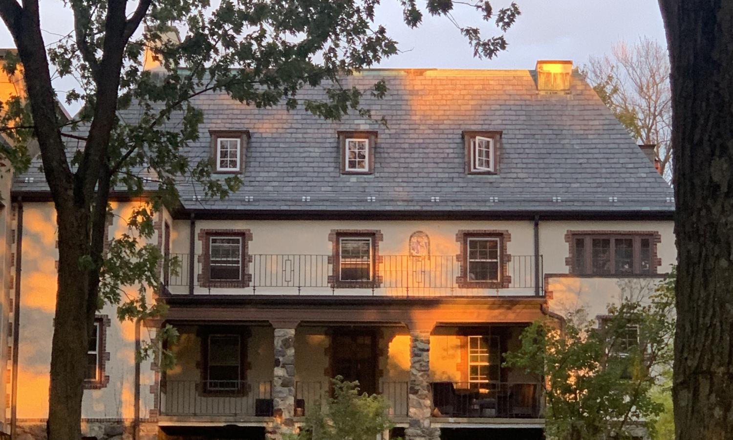 Bethany Arts Community main building at sunset. Large grass area in front of building with trees surrounding. Three floors with windows and porch seen on front of building.