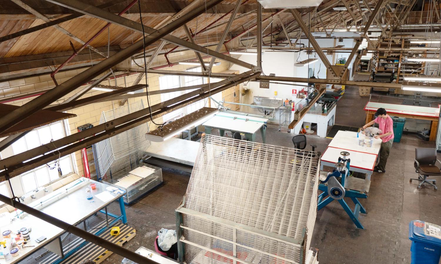 Light filled expansive room viewed from above containing rows of tables, drying racks, and printmaking equipment.  A person in a pink shirt is setting out containers of ink and preparing to work at one of the workstations.