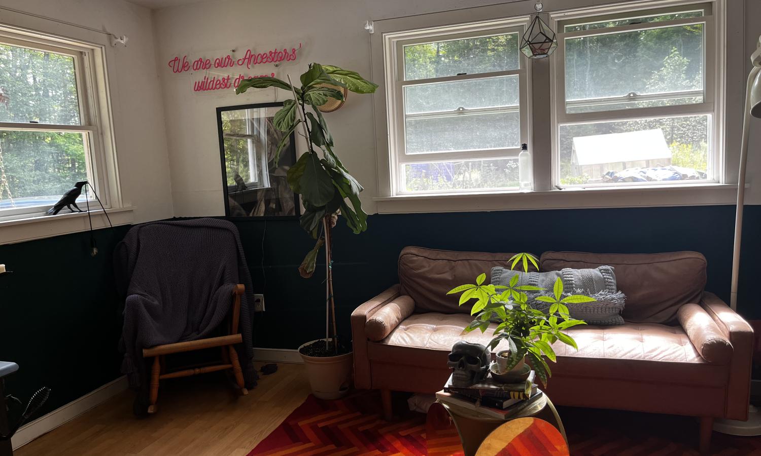 A brown, leather couch against a window with a neon sign beside it that reads "We are our Ancestor's wildest dreams" and a large fiddle leaf fig tree