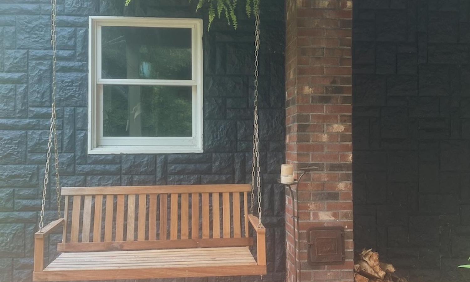 A wooden porch swing hangs outside a stone, dark-gray home with a painted concrete patio