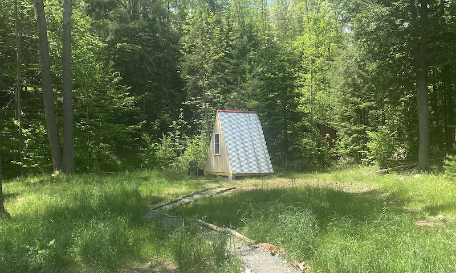 A small A-Frame structure sits on a field alongside a stream, in front of a forrest