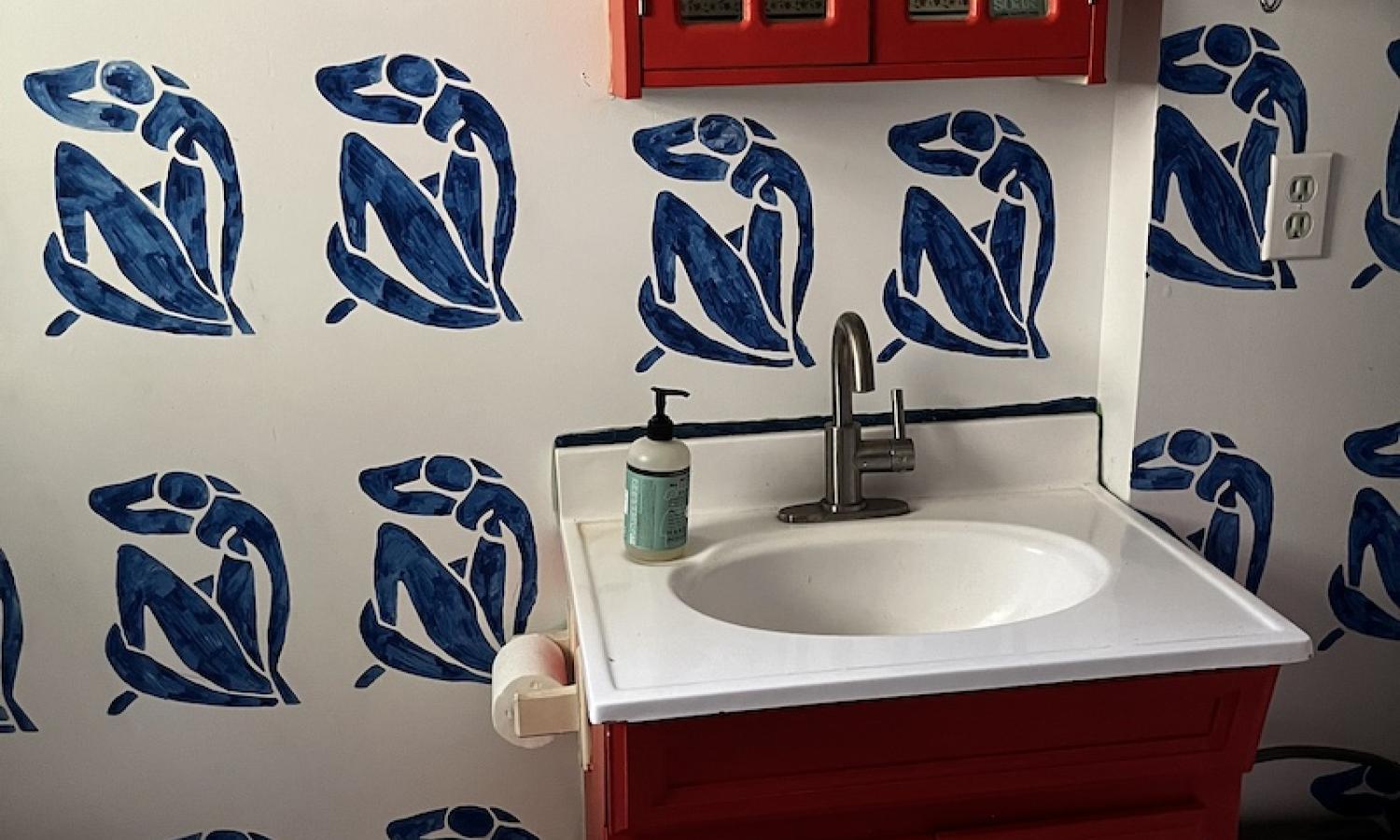 A toilet, sink, and medicine cabinet against white walls painted with blue figures. The sink vanity and medicine cabinet are dark orange