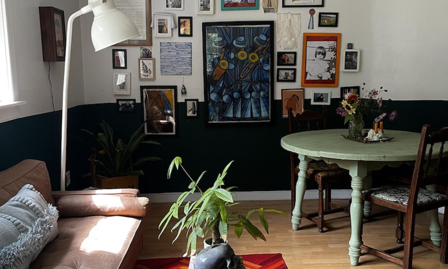 A living room with wood floors, an orange and red rug, a round sage-colored table, and a gallery wall of various photos and paintings