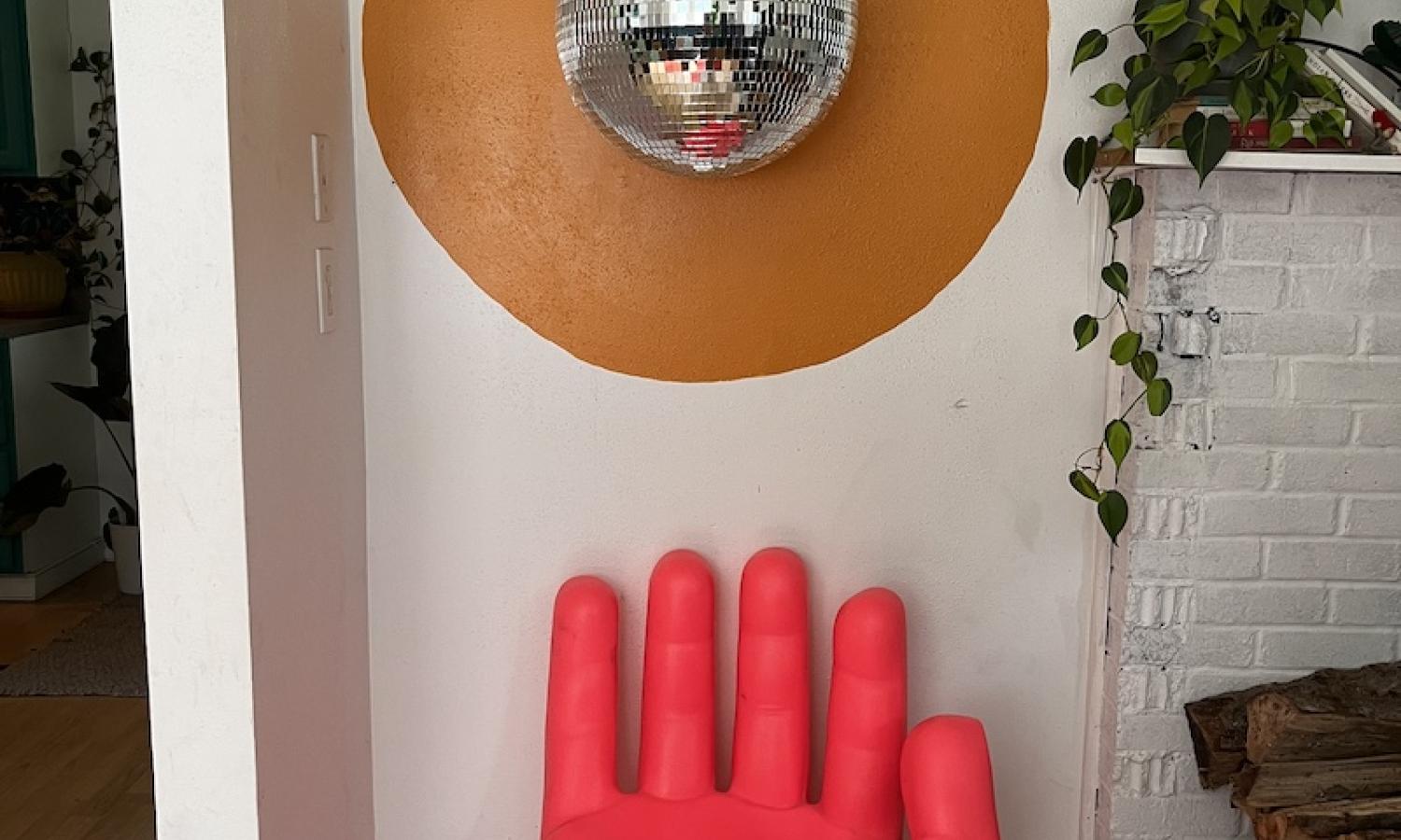 A hot-pink hand-shaped chair against a wall with a golden circle painted on it. A disco ball hangs in front of the golden circle