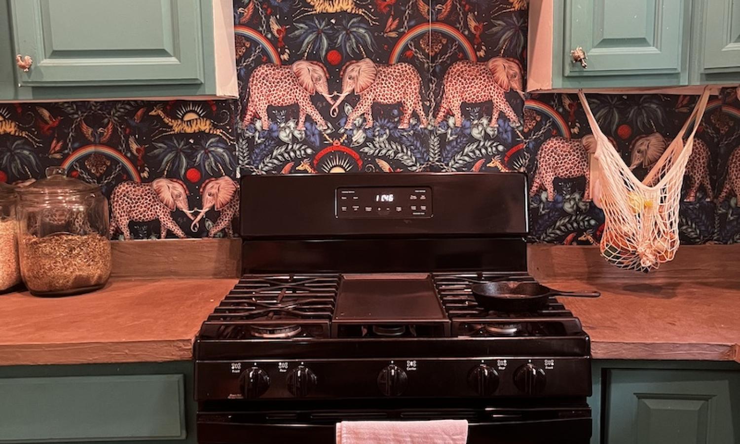 A black, gas over surrounded by teal cupboards and against a wall with mystical elephant wallpaper