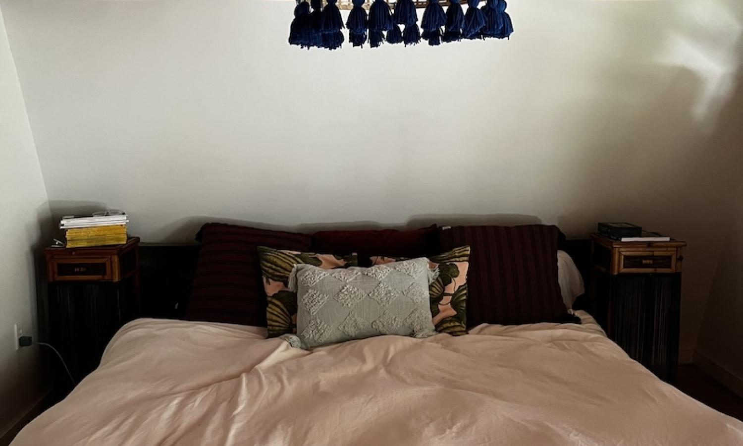 Large, king bed against a while wall with a coral colored ceiling and whicker lamp with blue tassels.