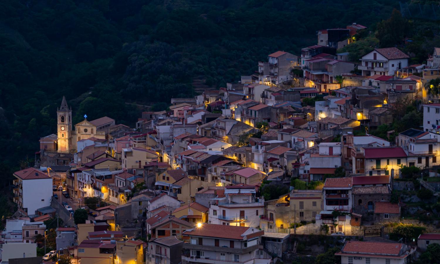 A view of the village of Pezzolo at night. The village is constructed on the side of a mountain, with a church steeple illuminated on the left side of the image. The houses are softly illuminated by streetlamps.