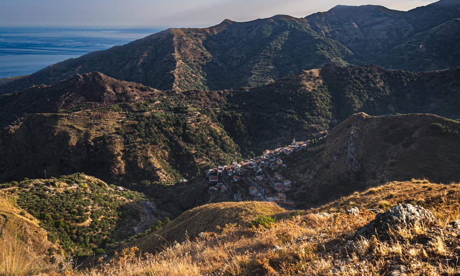 View of the village of Pezzolo from above. The village is nestled amongst mountains, with the sea visible in the background.