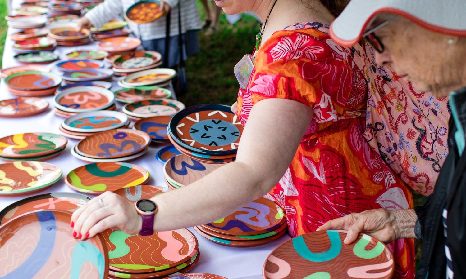 A woman in a red dress chooses one colorful plate from a table of bright, handmade ceramic plates.