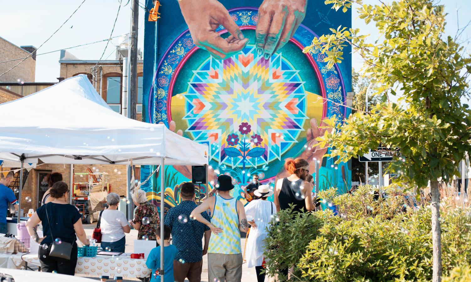 A colorful mural of hands and a star quilt on a tall building, with people facing it at an event.