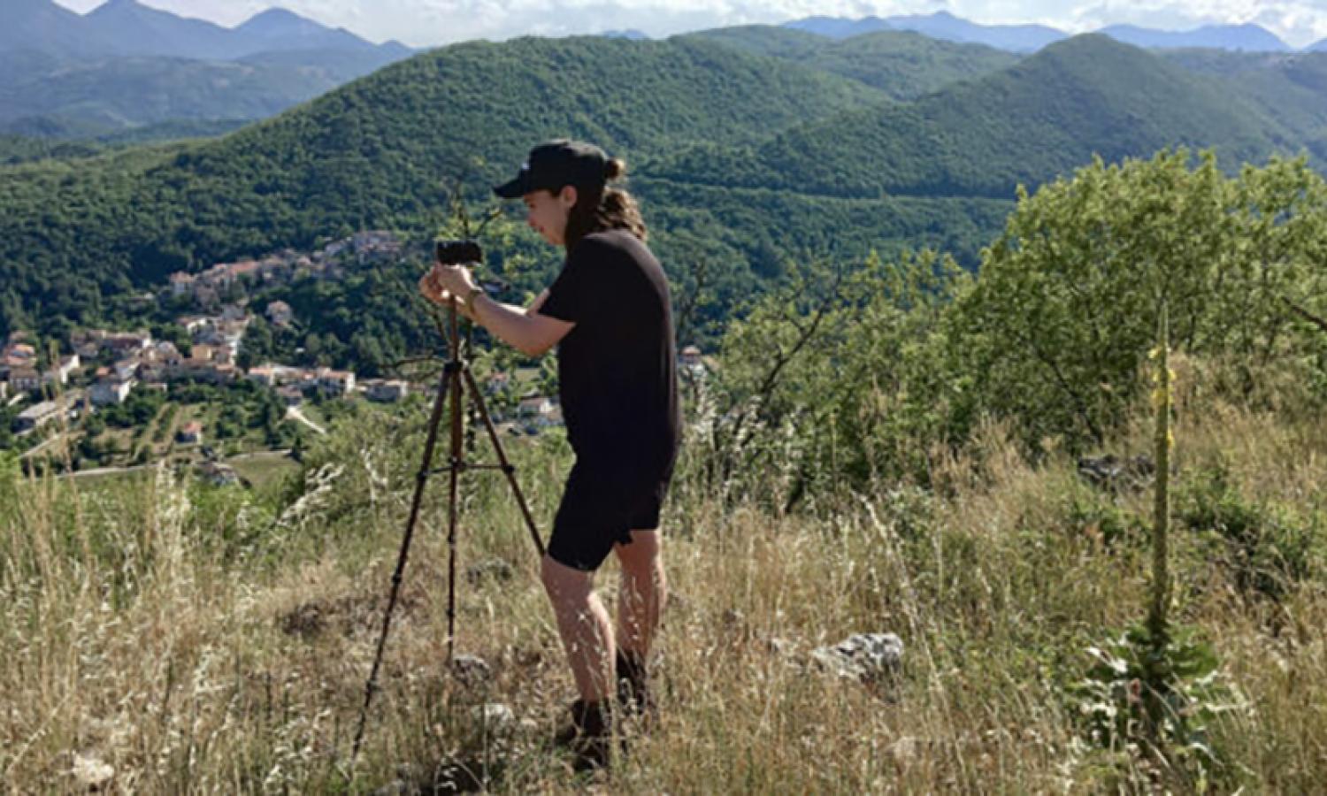 A woman wearing shorts stands with a tripod on a hillside overlooking mountains and a village