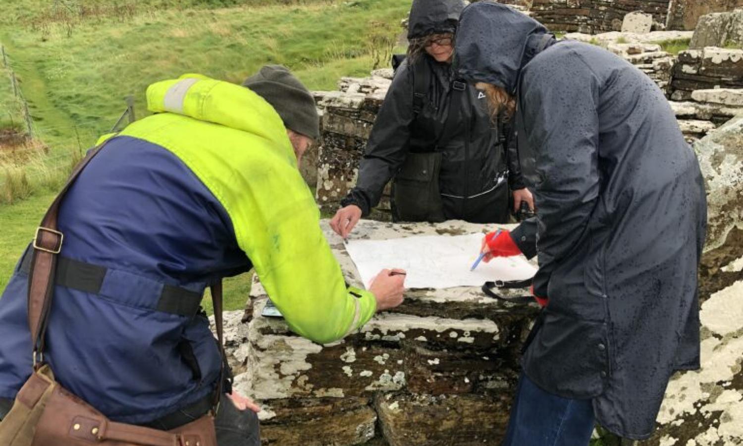 Three figures in wet weather clothing work together drawing on paper at an ancient site