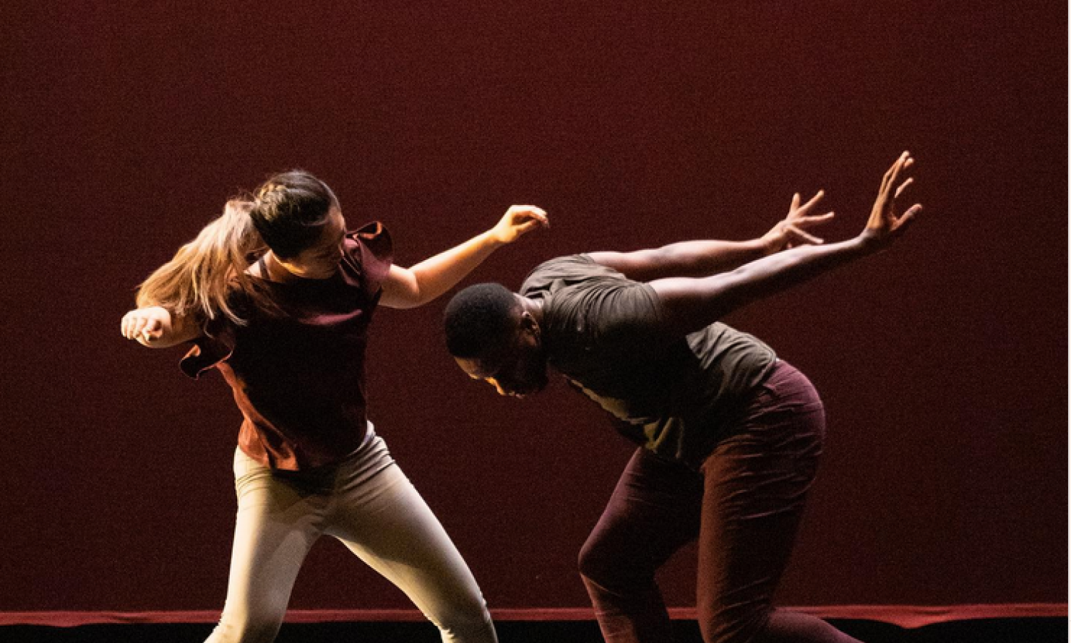 Two dancers wearing pedestrian-styled clothes are standing in a duet-pose. The person on the left appears braced for impact, with arms raised and legs slightly apart and bent. The person on the right is leaning forward from the hips as if to fly, with legs in a slight lunge and arms raised behind. The stage lighting is a dark red.