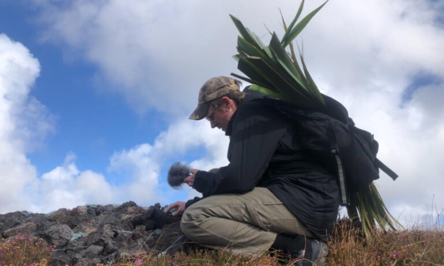 A figure crouches on rough ground with sky behind, they are collecting natural materials