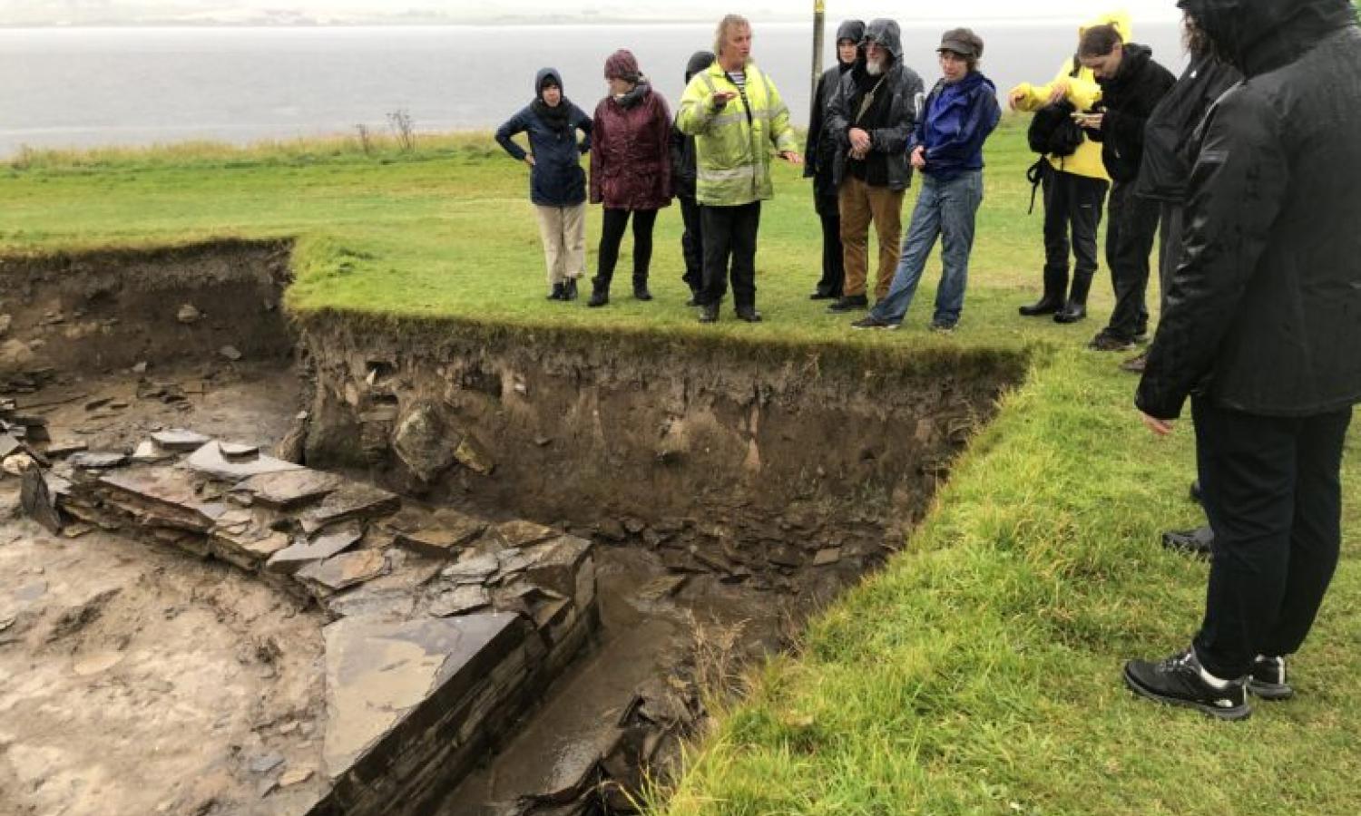 A group of people stand beside an open archeological trench