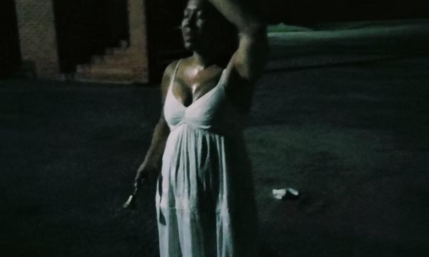 Still from site responsive performance Saltwater Road 2023. A woman wearing a white dress holding a bell in her right hand and left hand to head in the dark of night with a single light from off screen.