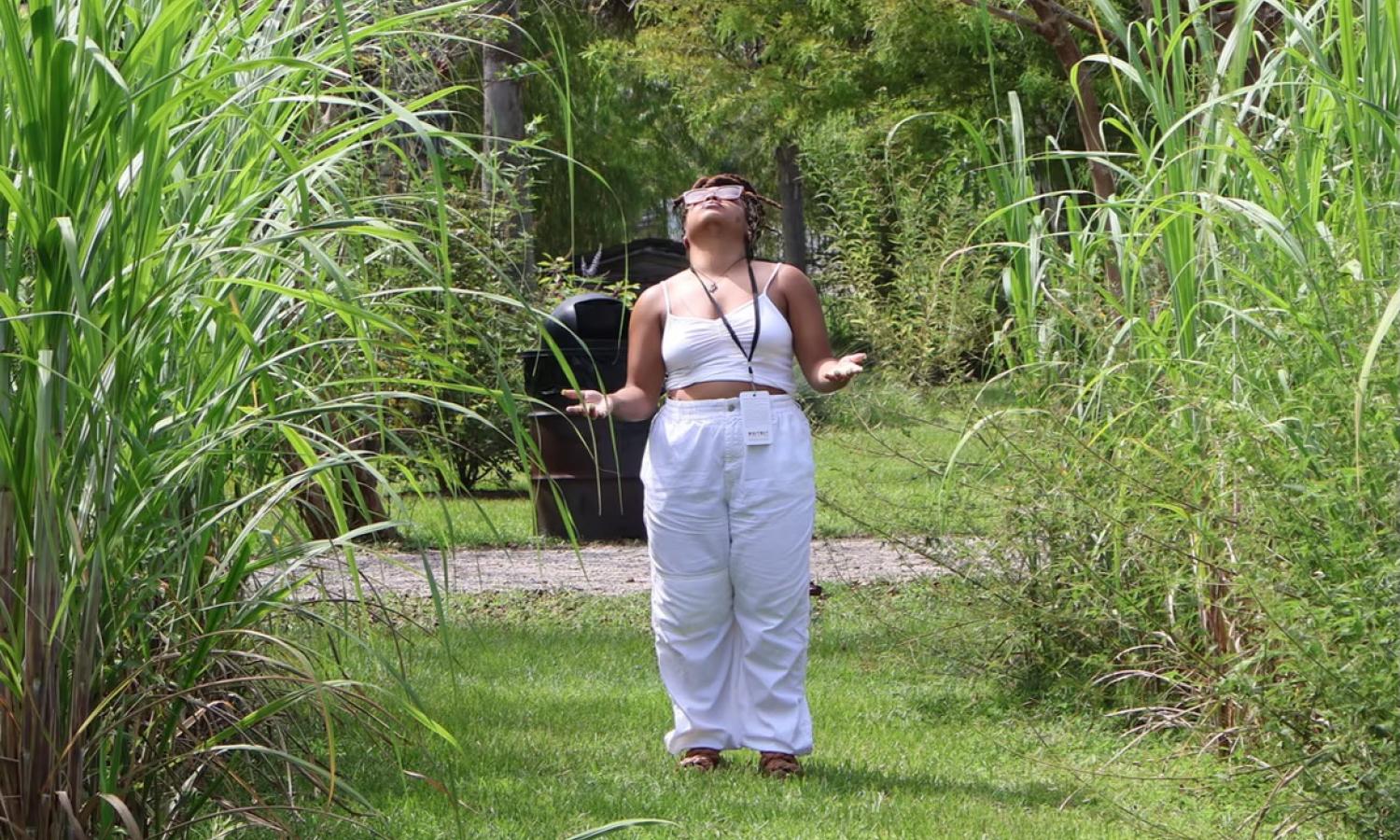 Still from site responsive performance Saltwater Road 2025. A woman wearing white with hands open, eyes closed, looking up, in the day, on a grassy path, between tall reeds of grass.