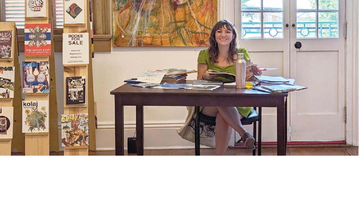 Bethany Mulligan, Kolaj Institute volunteer and nearby contact, sits at a studio desk surrounded by Kolaj Magazine displays, artwork, and balcony doors.