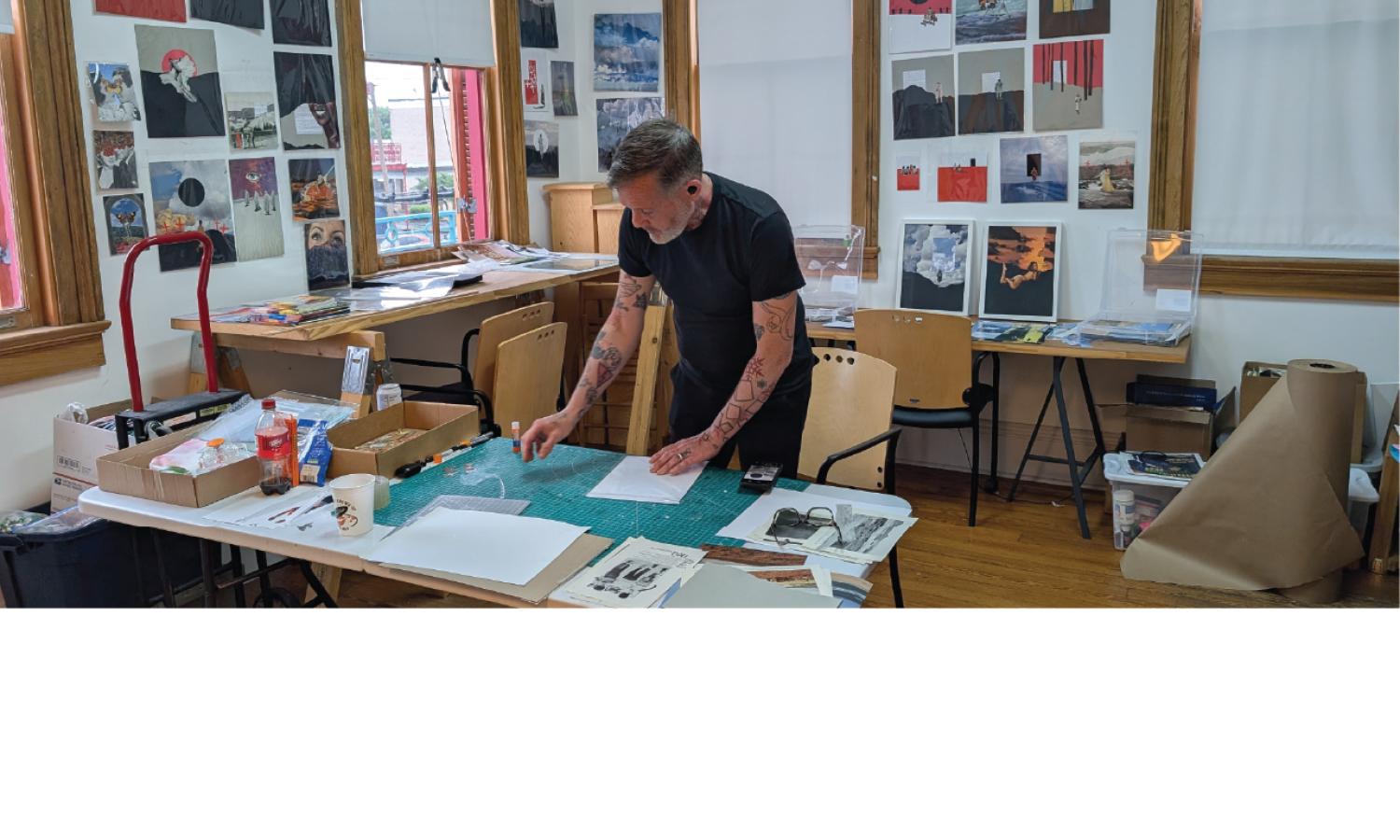 Resident artist Levi Eleven works at a studio table, surrounded by collage materials, tools, papers, and works-in-progress.