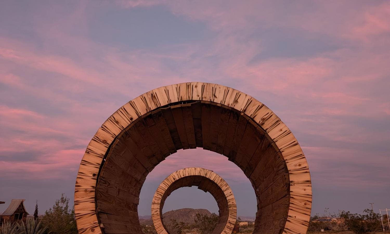 Wooden circular sculptures in a field with colorful sunset in sky.