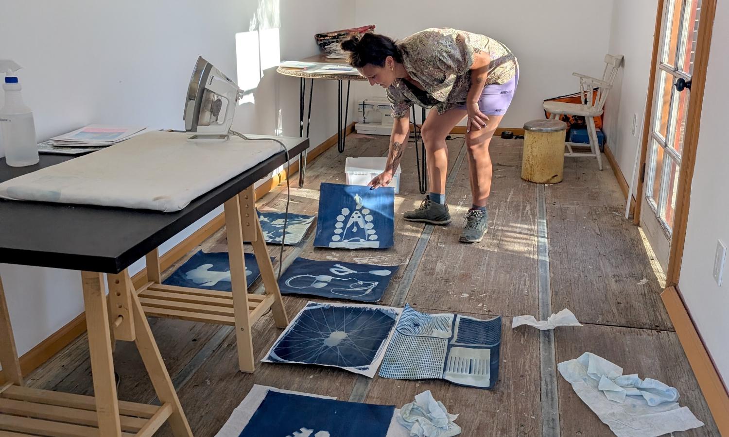 Image shows an artist at work in side container studio, with white walls, wood floor and art work spread out on floor.