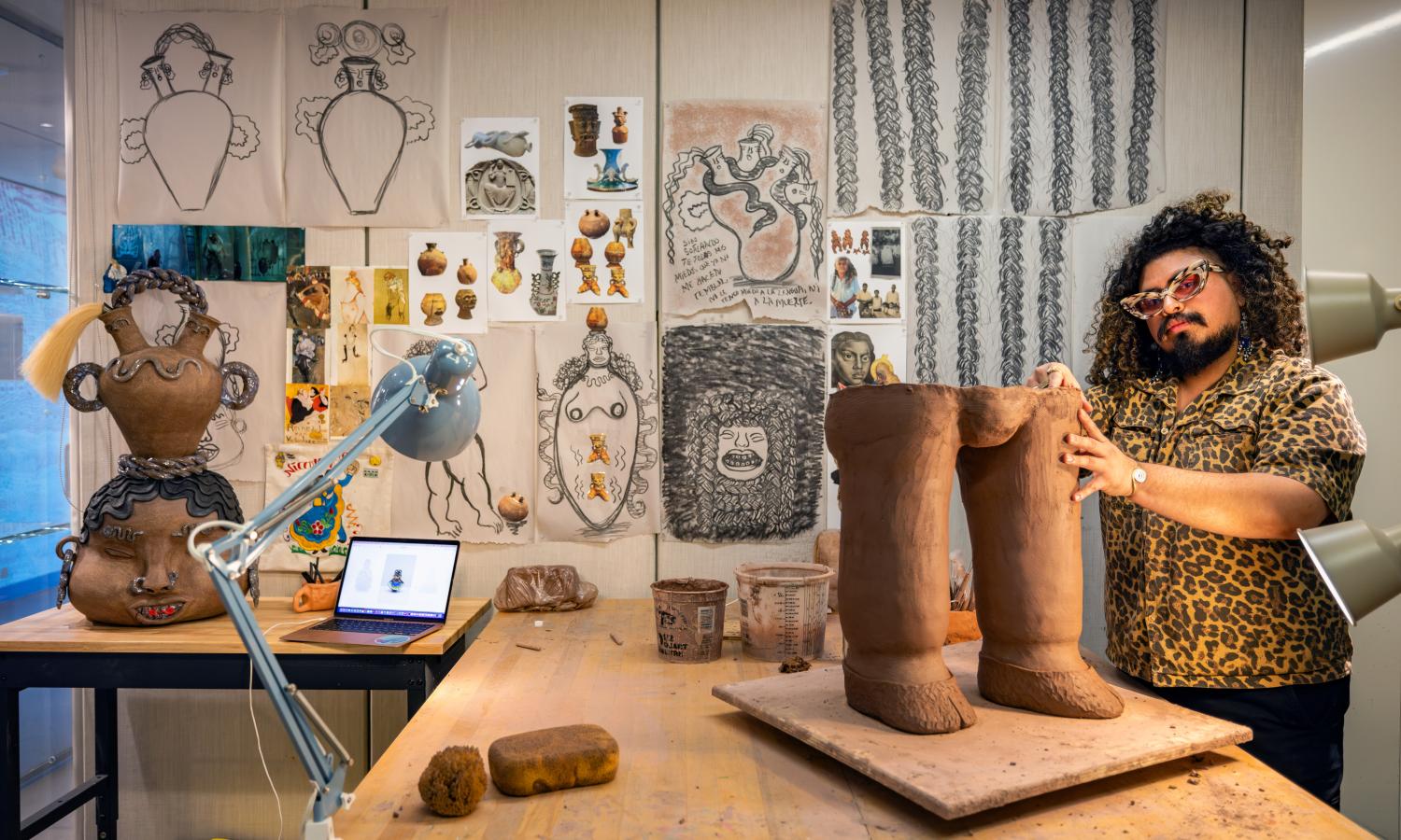 An artist wearing sunglasses and a leopard shirt with dark curly hair works on a clay sculpture in his studio