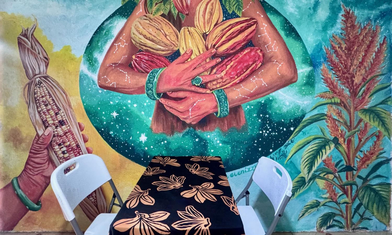 A mural of a person holding cacao pods in their arm. On the left, an elderly hand holds corn. To the right is an amaranth plant.
