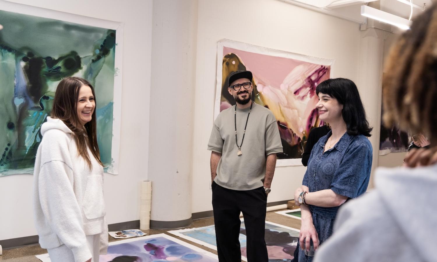 view of a studio visit in high ceilinged room with large paintings on the wall behind the female artist who is speaking with two other artists