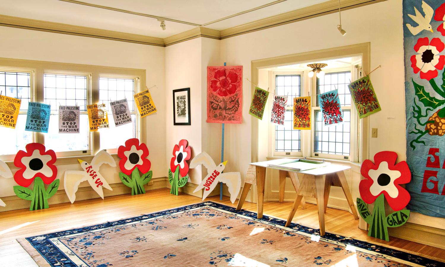 A white walled room with colorful prints hung on strings in front of them. A picket sign leans against the wall in the center of the image. A pink rug with a blue frame is on the ground. The edge of a large banner peaks out of the right.
