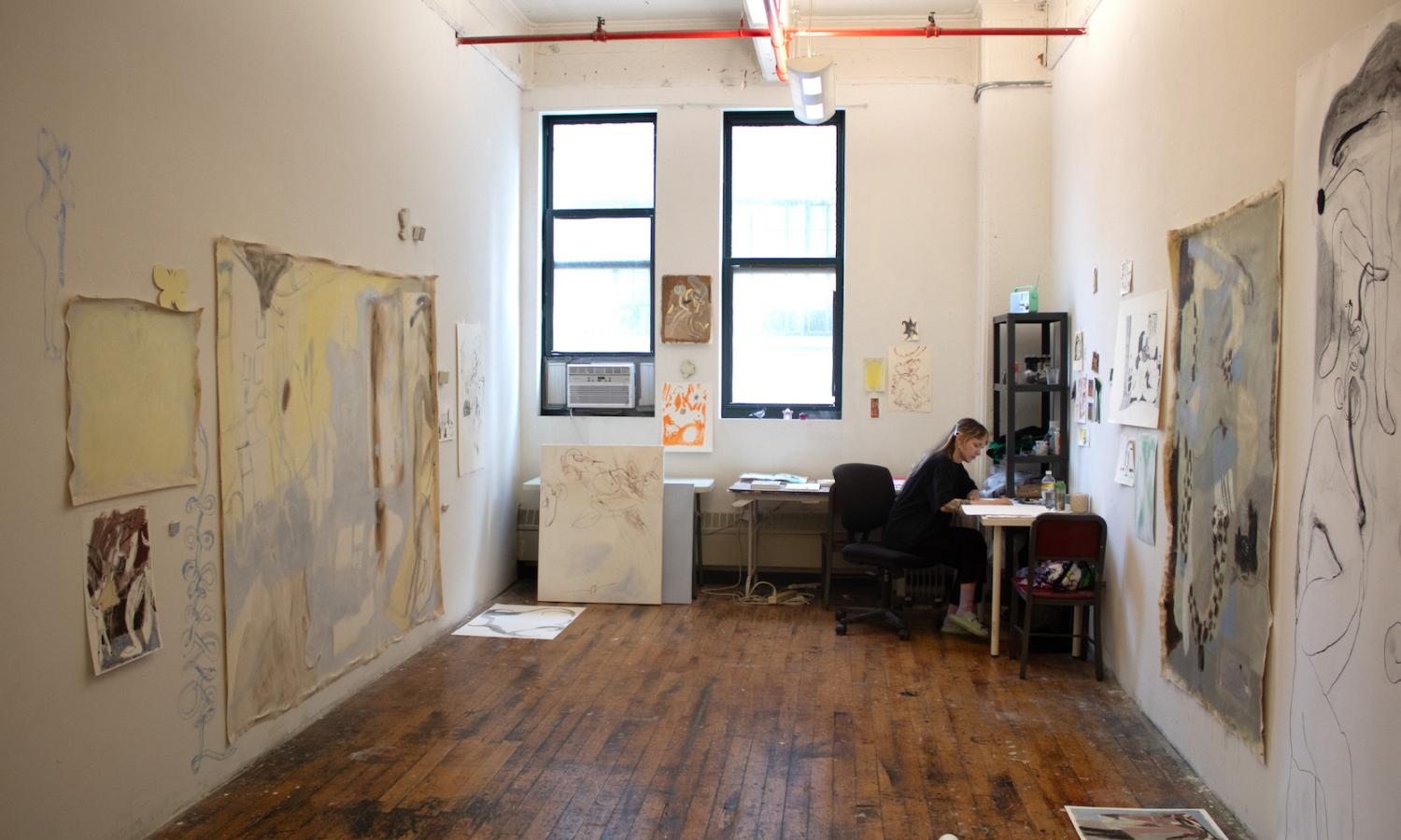 Image of artist sitting at a desk in the corner of a large private studio room, with paintings in progress surrounding them.