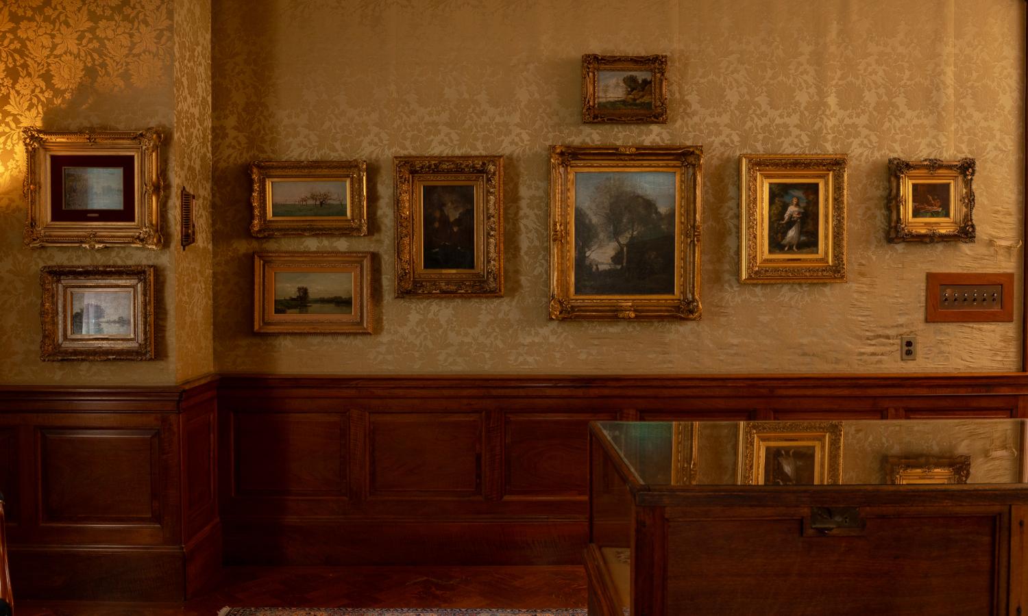 Gold silk damask covered wall is adorned with guilded framed oil paintings. The lower half of the wall is light wood paneled.
