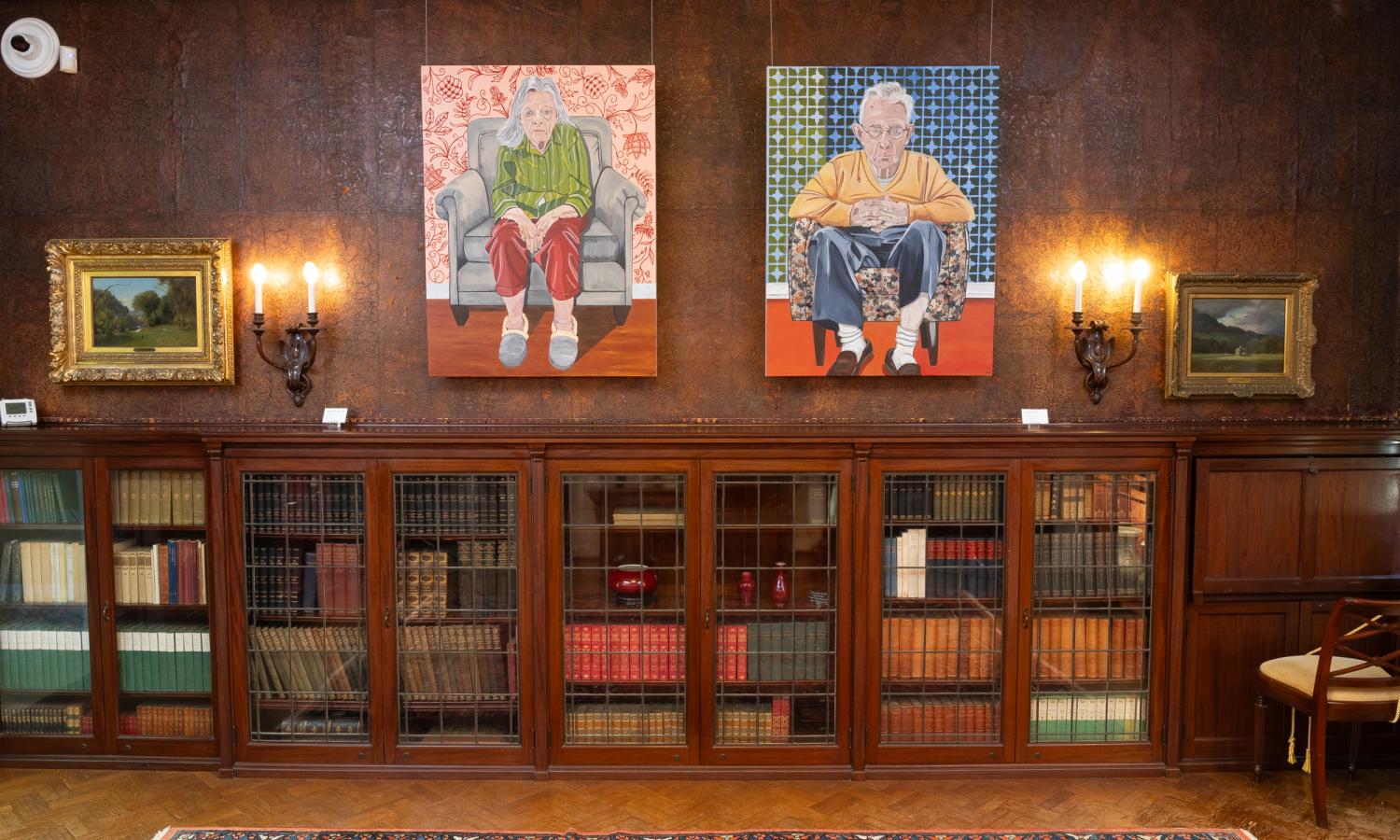 Two modern acrylic paintings on stretched canvas of elderly white people between two wooden sconces against leather wallcovering. Underneath are 3  built in bookshelves.
