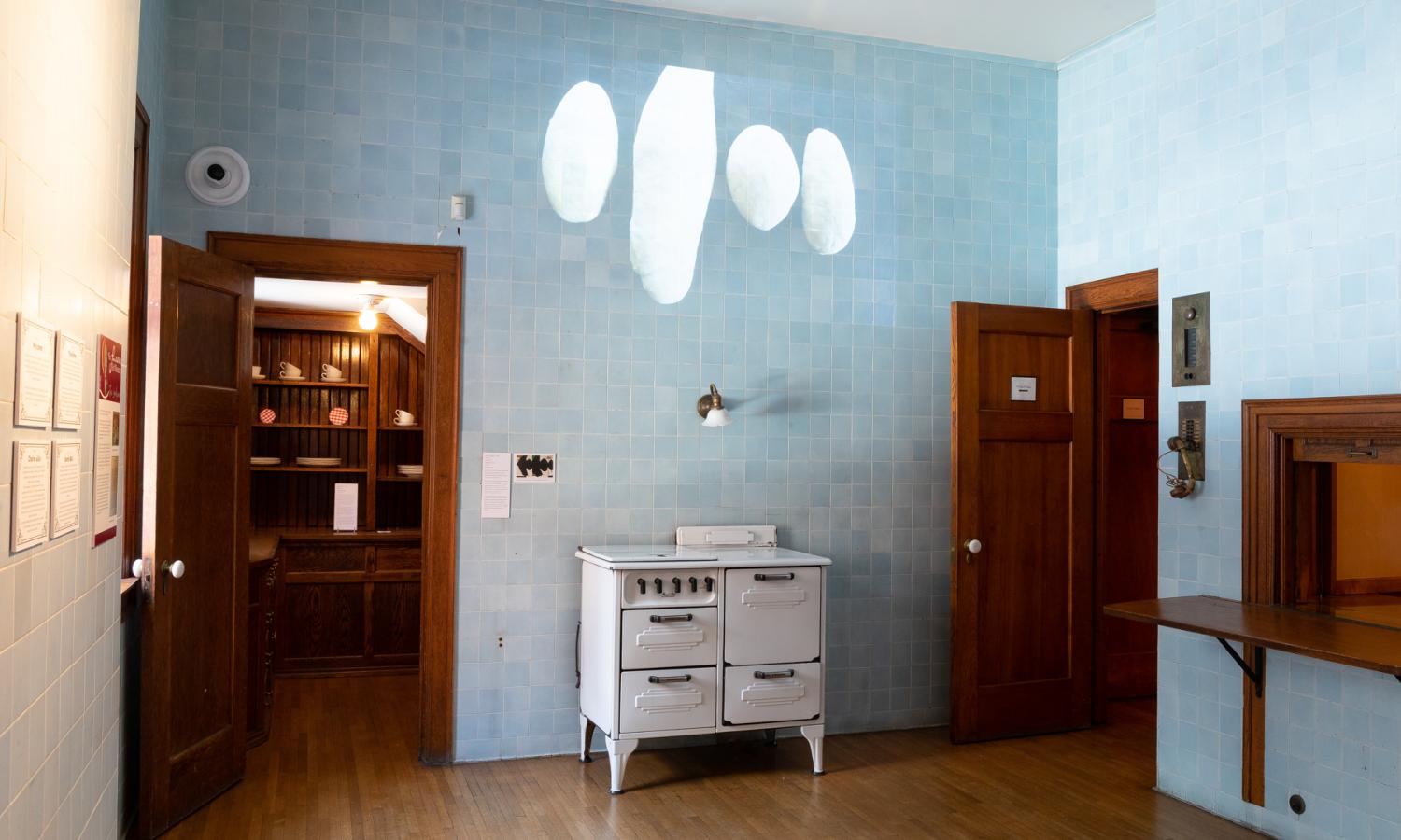 A projection above a white stove is shown against a blue tiled wall. An open door on the left shows a small walk in wooden shelved pantry. On the right shows an open wood door, a small phone system built into the wall, and a serving station passway framed in wood.