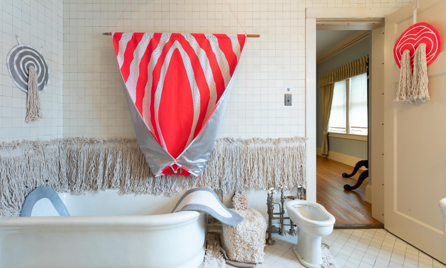 A tub and bidet  are shown in the foreground against a white tile wall with a pink textile sculpture in a triangle shape comes down off the wall into the bath tub. An open door on the right shows the windows of the other room.